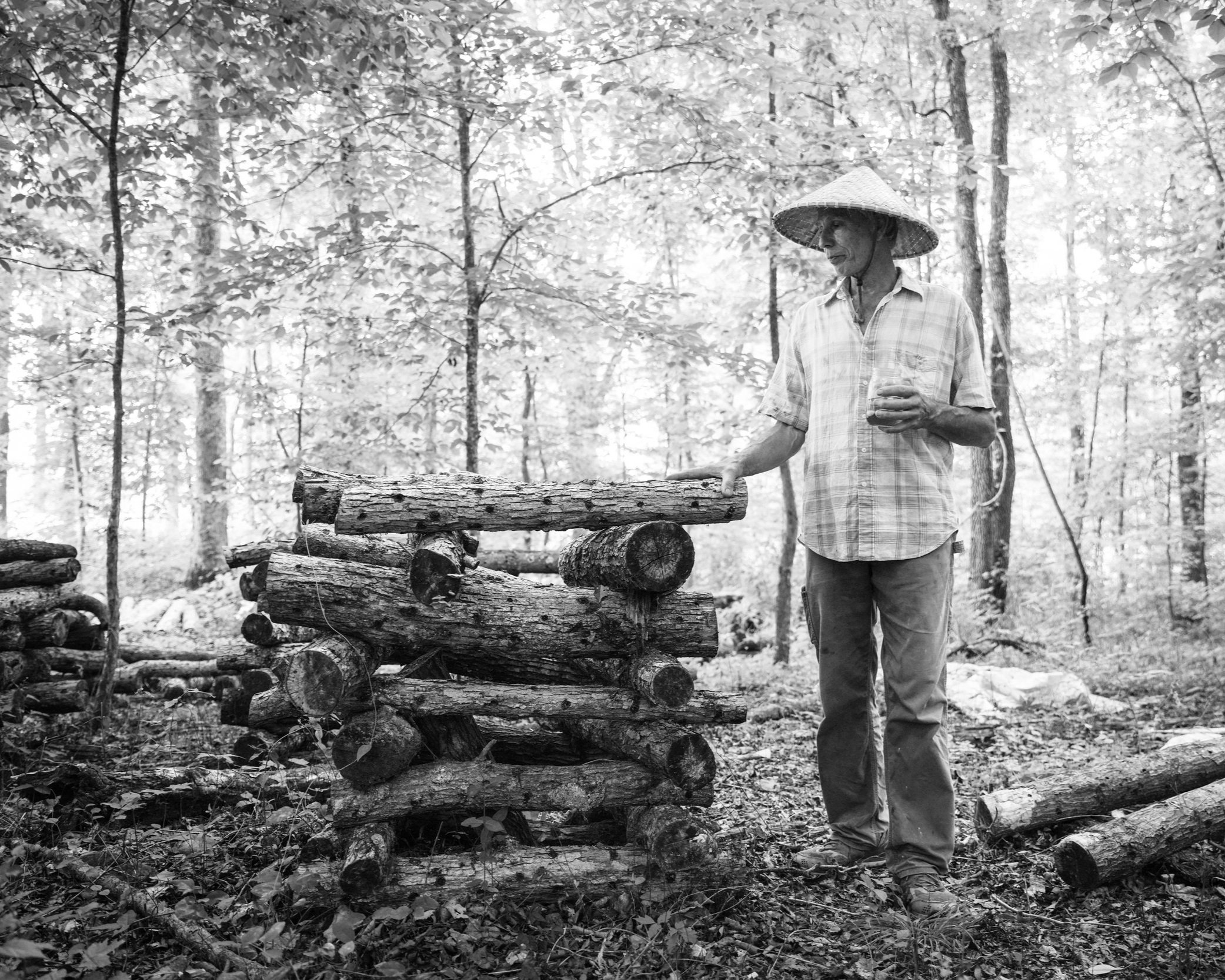 2022_sequatchie+cove+farm_rinne+allen-09613 Man in a straw hat stands in a forest, arranging stacked logs while holding a drink, surrounded by trees in a serene, black-and-white setting.