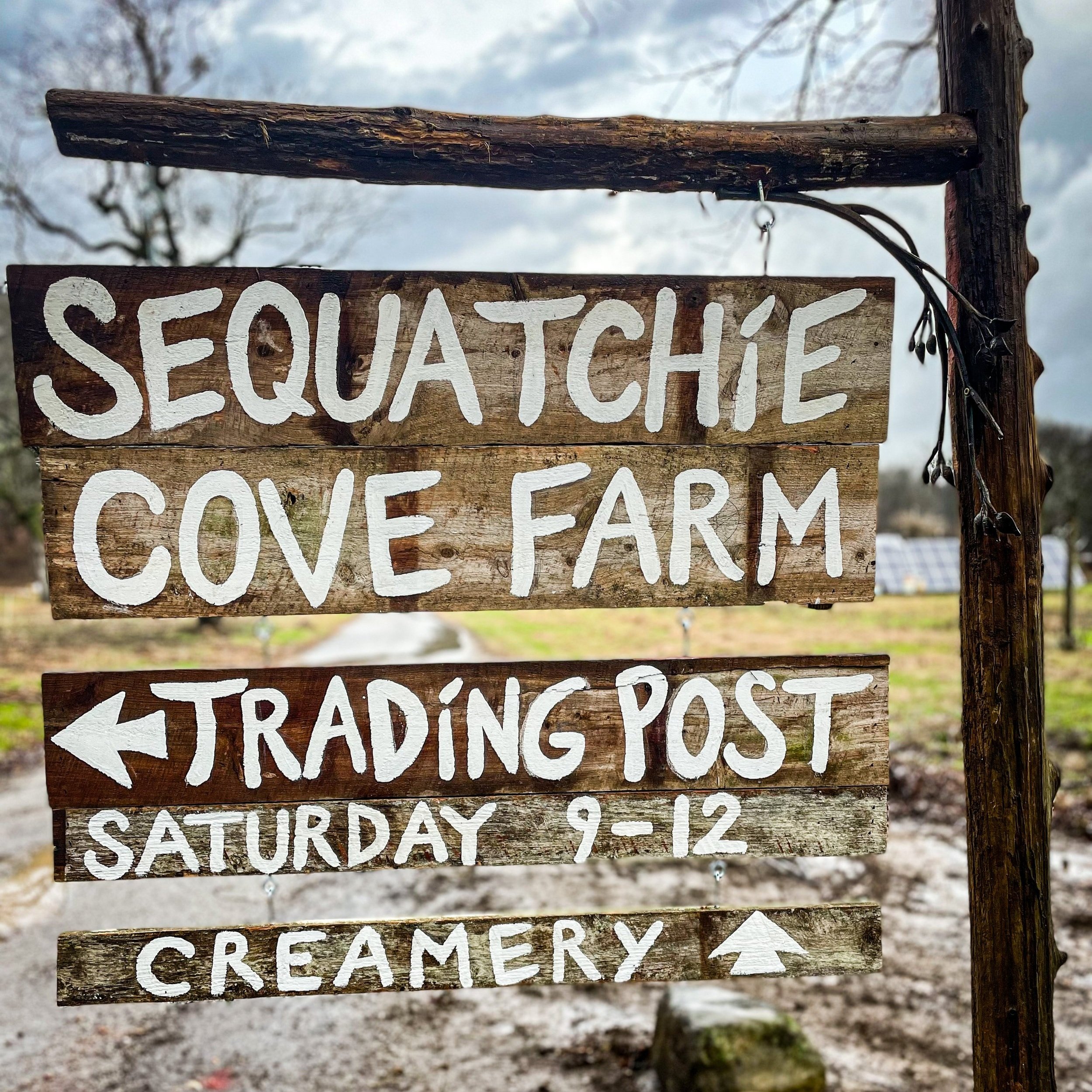IMG_2726_jpg Sign for Sequatchie Cove Farm, featuring directions to the Trading Post and Creamery, with trading hours on Saturdays from 9 AM to 12 PM. The background shows a rural landscape with solar panels.