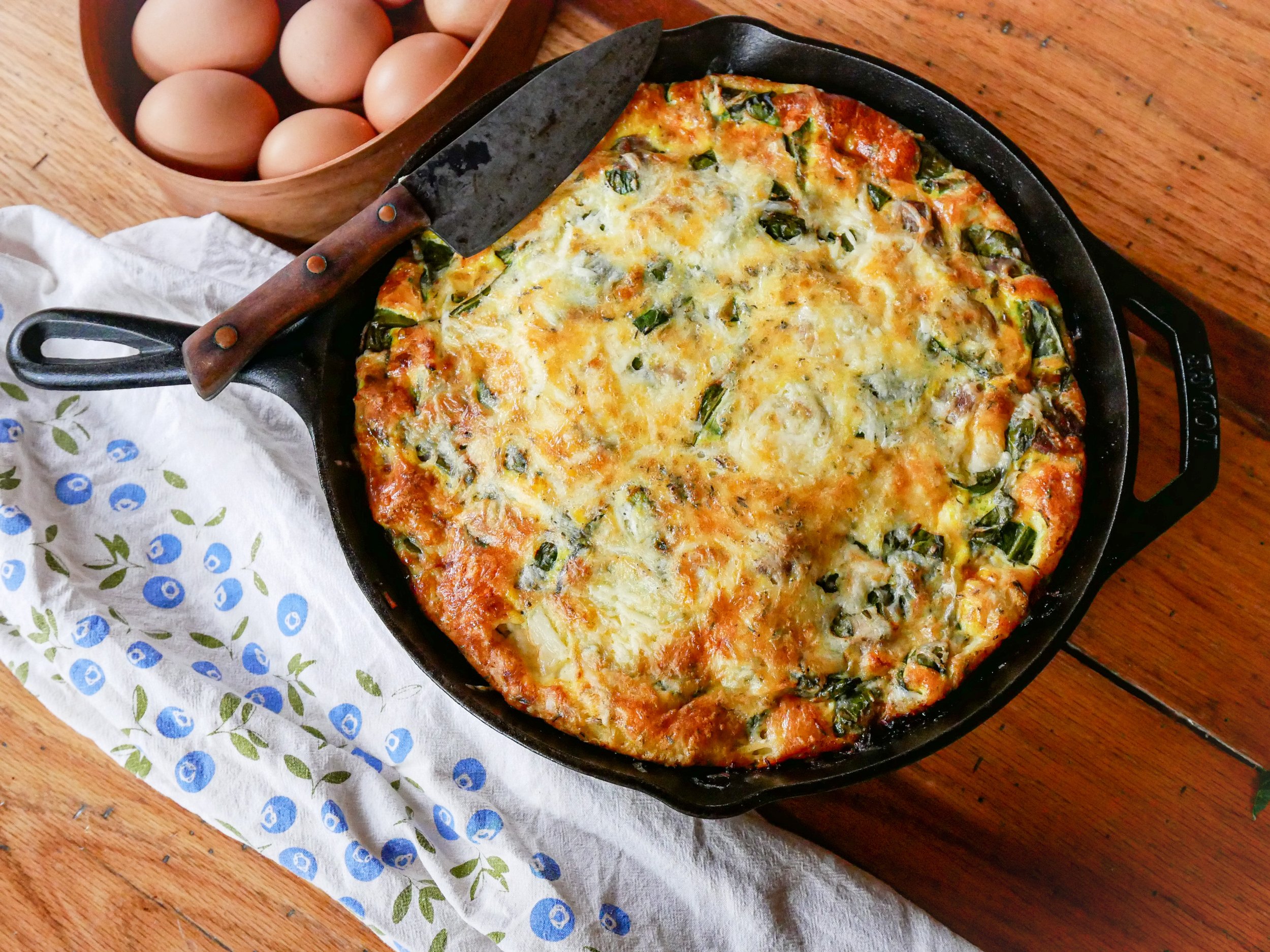 Cast iron skillet with a golden-brown frittata made with fresh greens, placed on a wooden table next to a bowl of eggs and a floral kitchen towel.