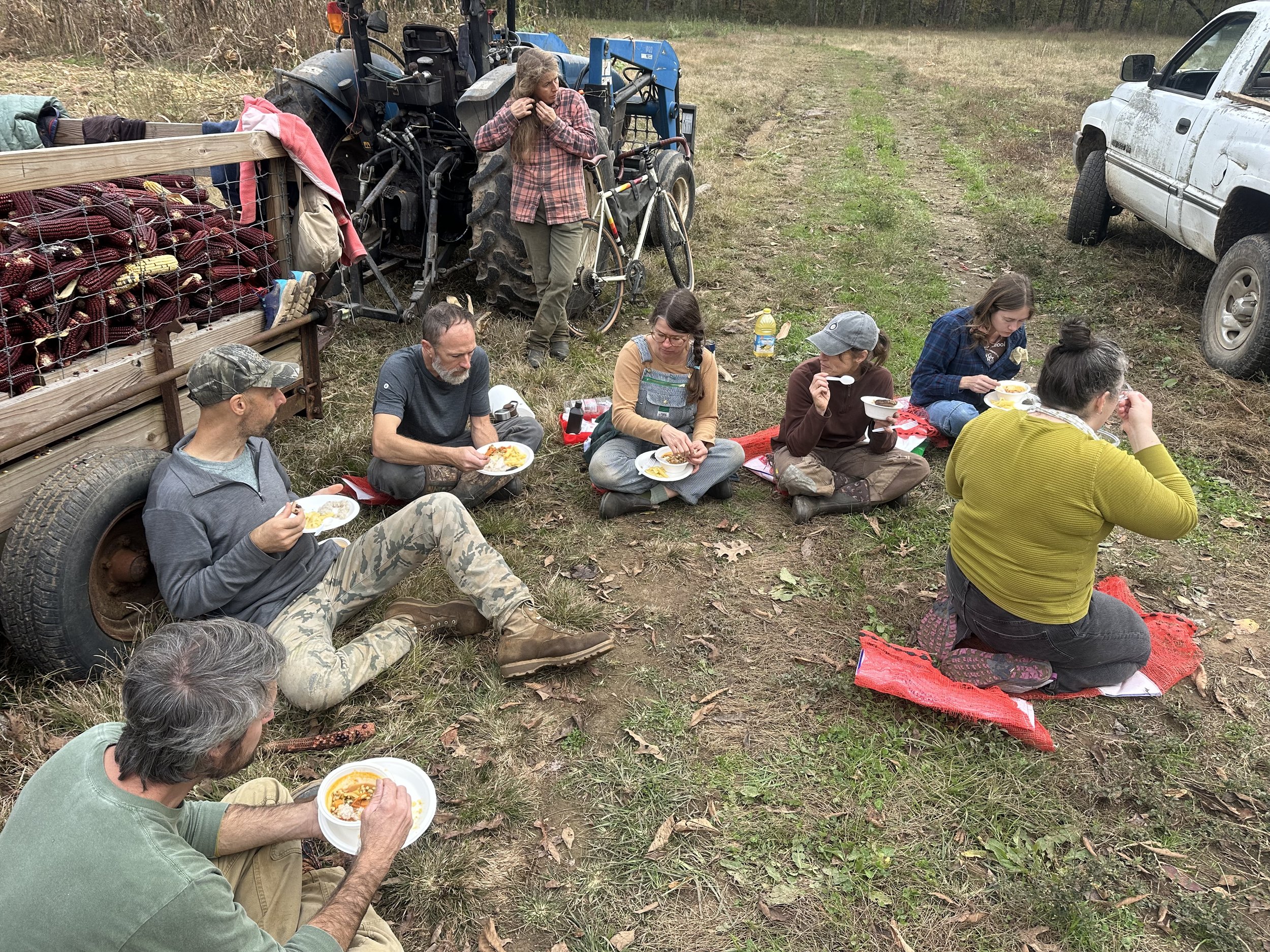 Group of eight people enjoying a meal outdoors on a farm, seated on red mats near a tractor and a pickup truck, with a trailer loaded with harvested produce in the background.