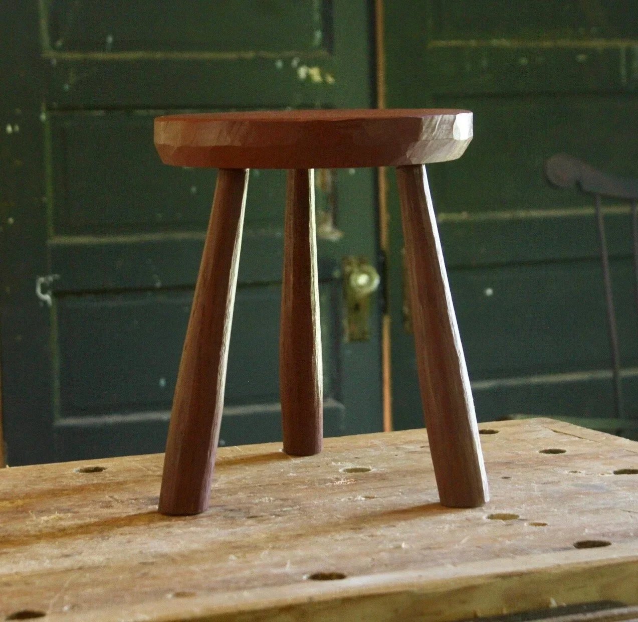 Wooden stool with a round top and three tapered legs, resting on a workbench in a rustic workshop setting. The background features dark green walls and a vintage door, highlighting the craftsmanship of the stool.
