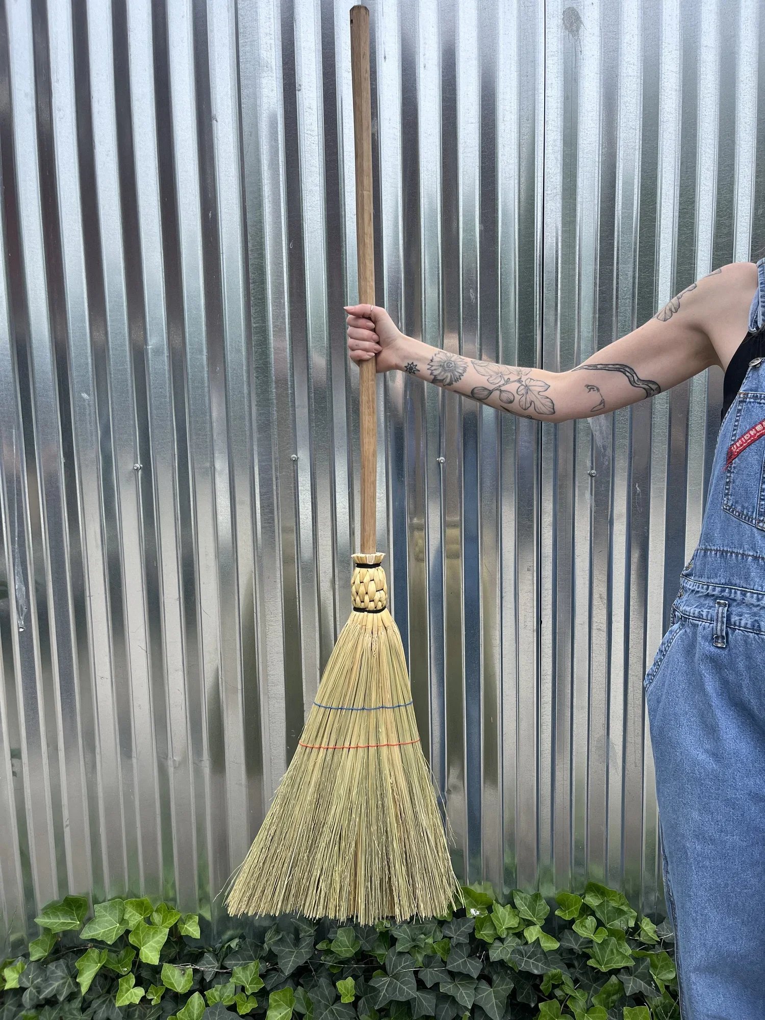 Hand holding a traditional straw broom with a wooden handle, set against a corrugated metal background and surrounded by green ivy, showcasing a blend of rustic charm and functionality.