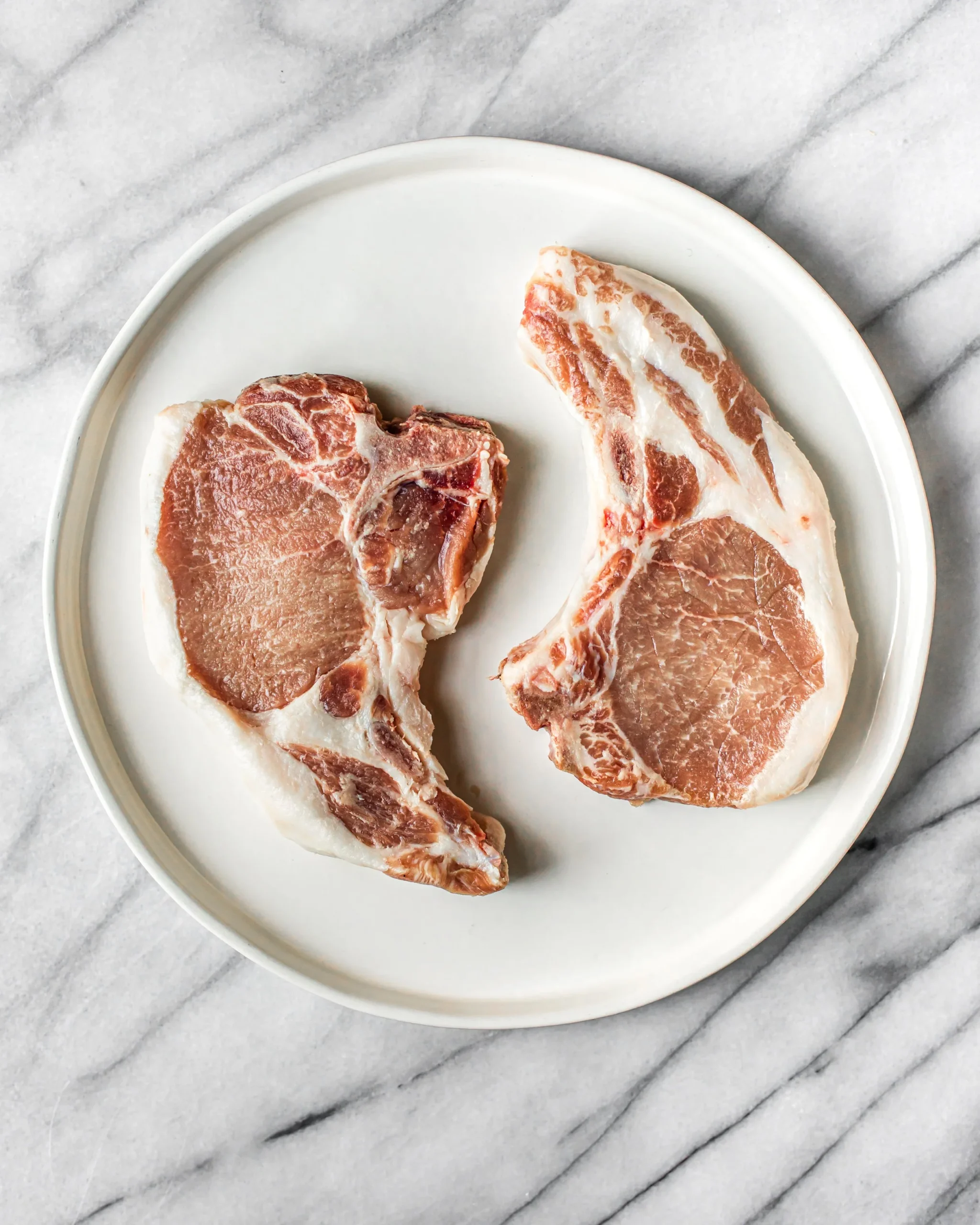 SCFporkchops-2 Two raw pork chops on a white plate, showcasing marbling and pink meat against a light marble countertop background, ideal for recipes or cooking tips.
