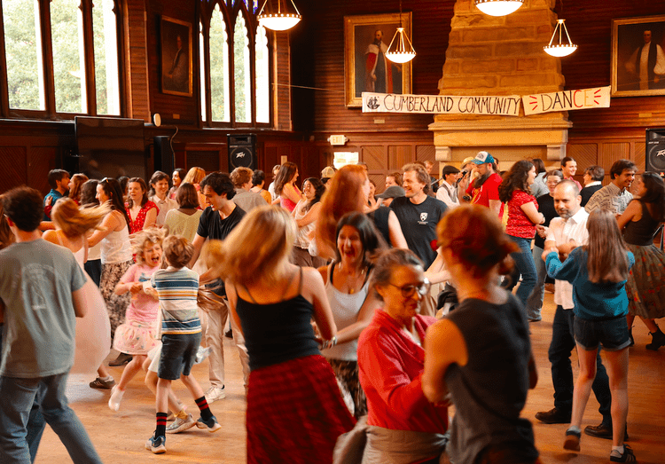 A lively community dance event at the Cumberland Community Center, featuring a diverse group of participants joyfully dancing together in a spacious hall adorned with historical portraits and warm lighting.