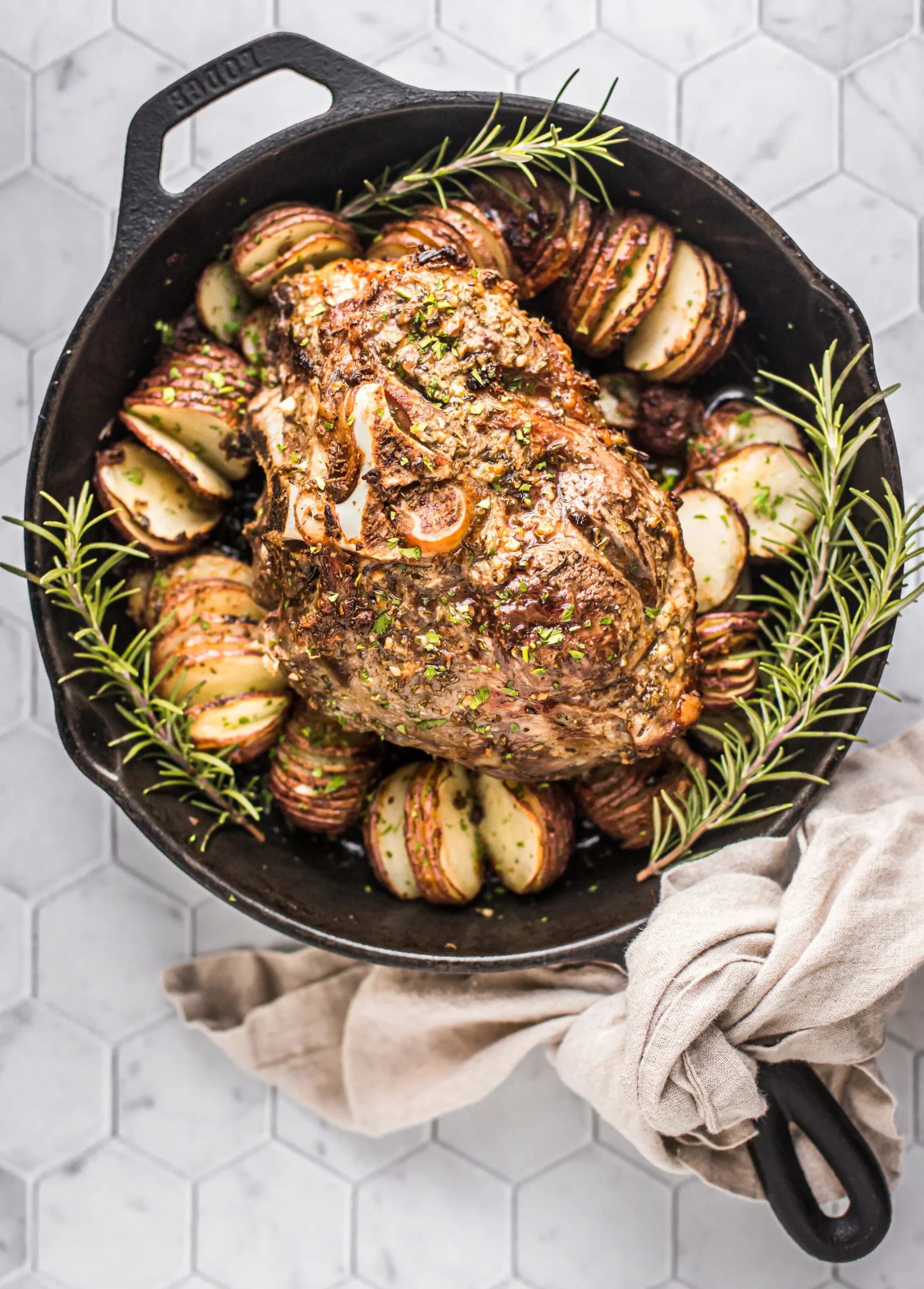 A succulent roasted lamb shoulder garnished with herbs, surrounded by sliced roasted potatoes in a cast iron skillet, showcasing a delicious and hearty meal.