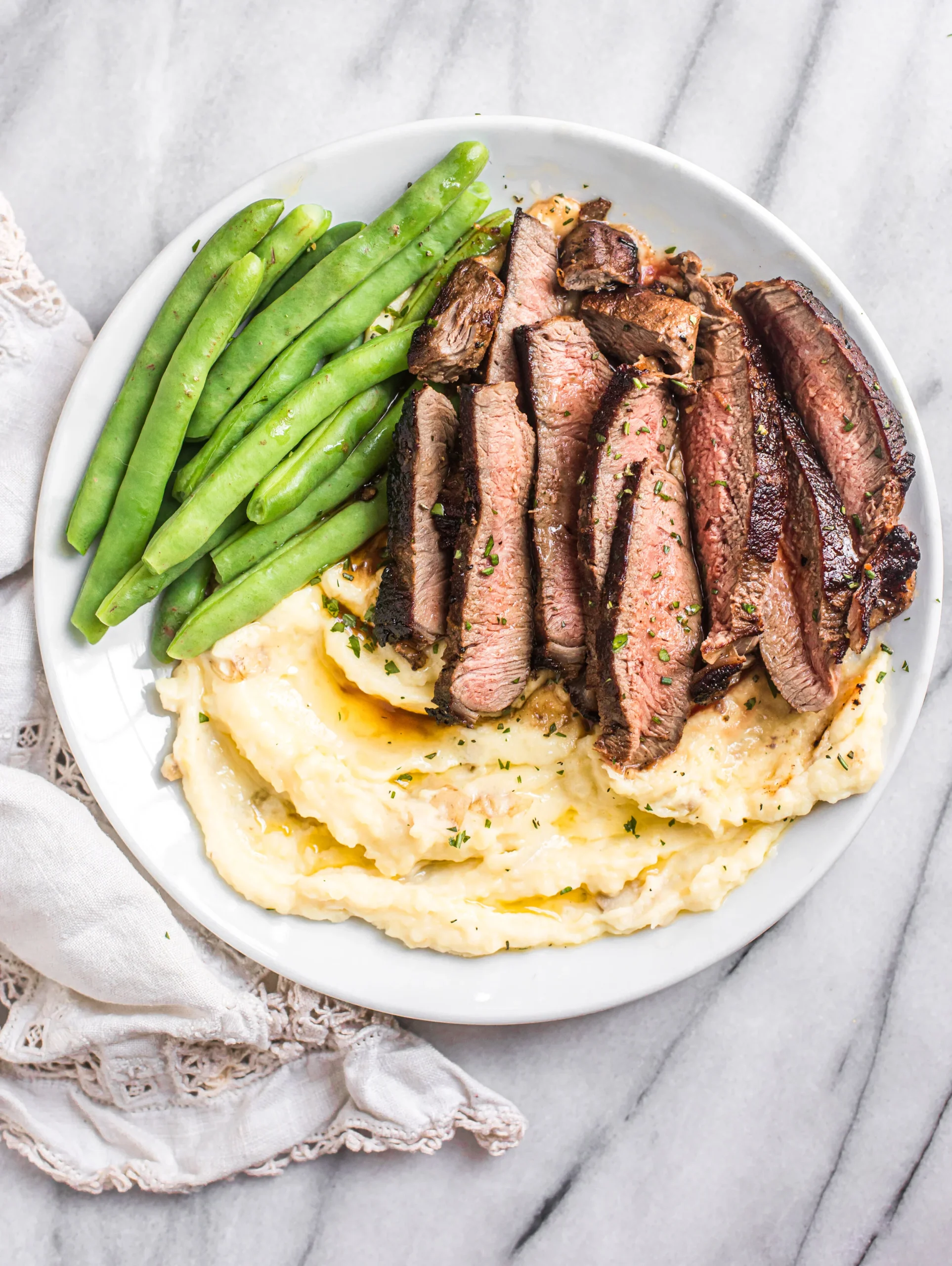 Plate of sliced steak served with creamy mashed potatoes and green beans, garnished with herbs, on a marble surface. Perfect for a hearty dinner.