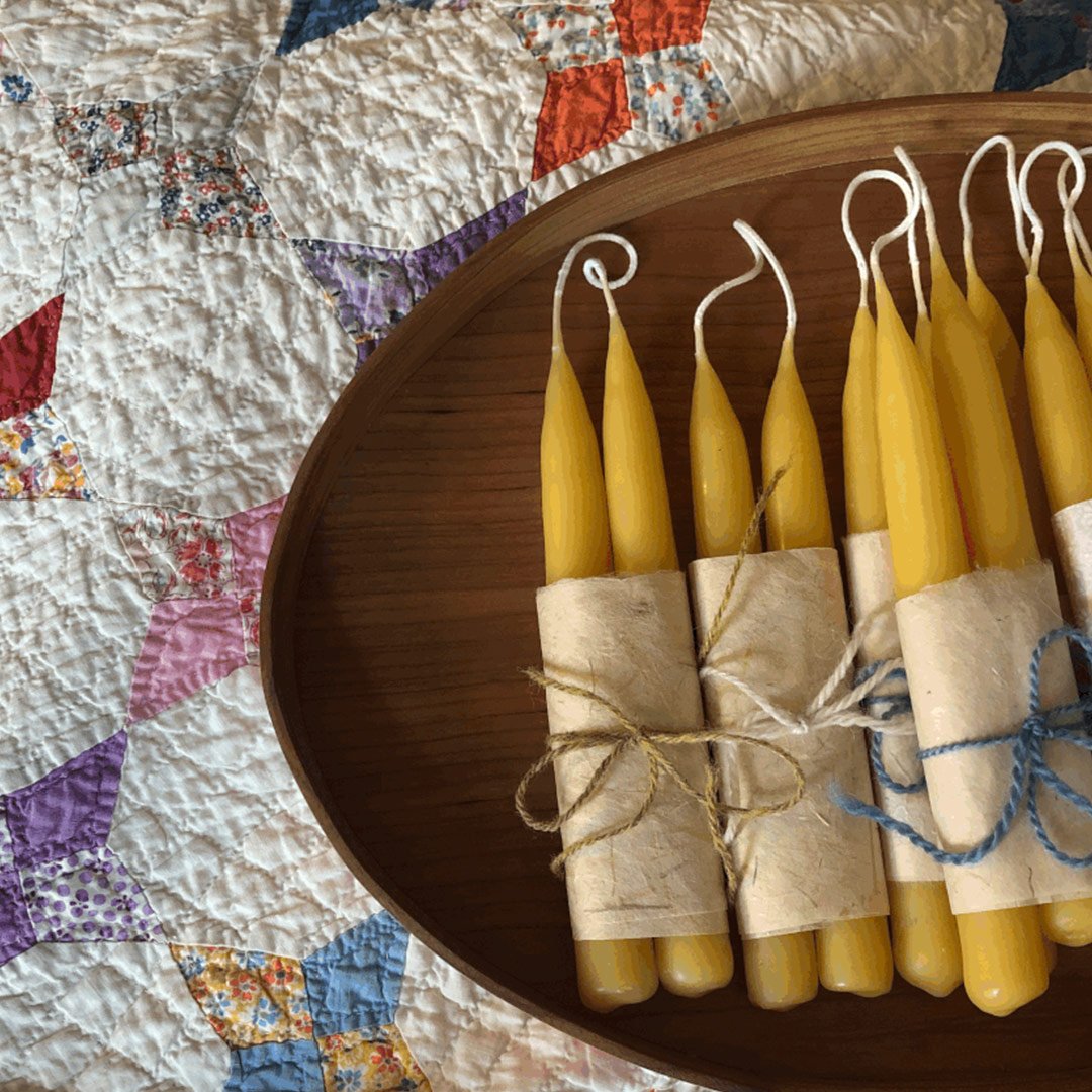 Handmade yellow taper candles bundled with natural twine, displayed on a wooden tray, against a colorful quilted background. Perfect for home decor or special occasions.