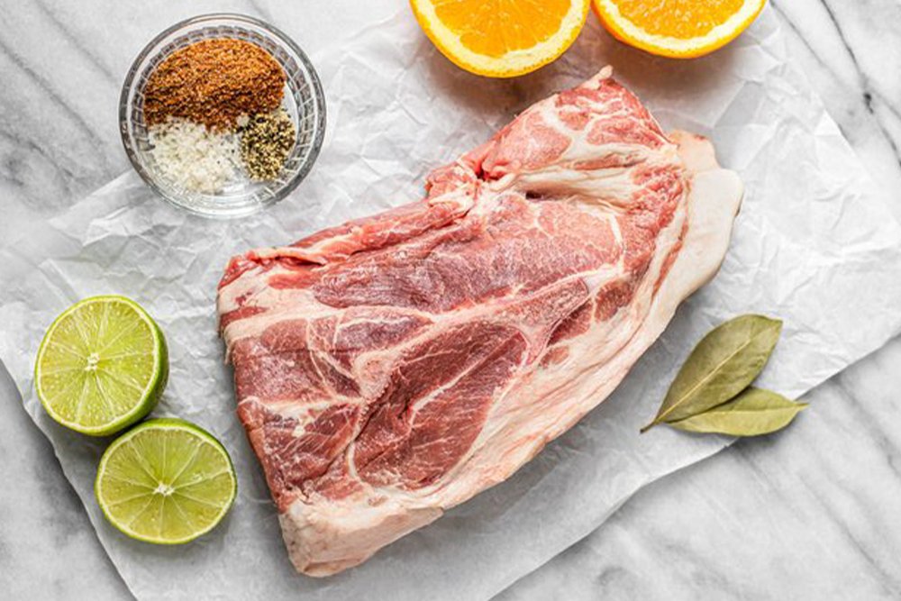 sequatchie.cove.farm.tennessee.heritage.pork.shoulder Pork shoulder cut on parchment paper, surrounded by fresh lime halves, orange slices, and a small bowl of spices including salt, pepper, and seasoning mix, ideal for marinating and cooking.