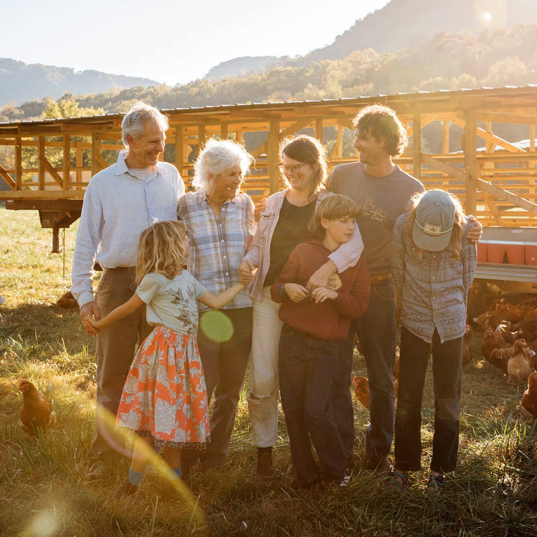 A diverse family of seven stands together in a sunlit farm setting, surrounded by chickens and a wooden chicken coop. The group, including children and adults, enjoys a moment of connection and laughter against a backdrop of rolling hills and trees.