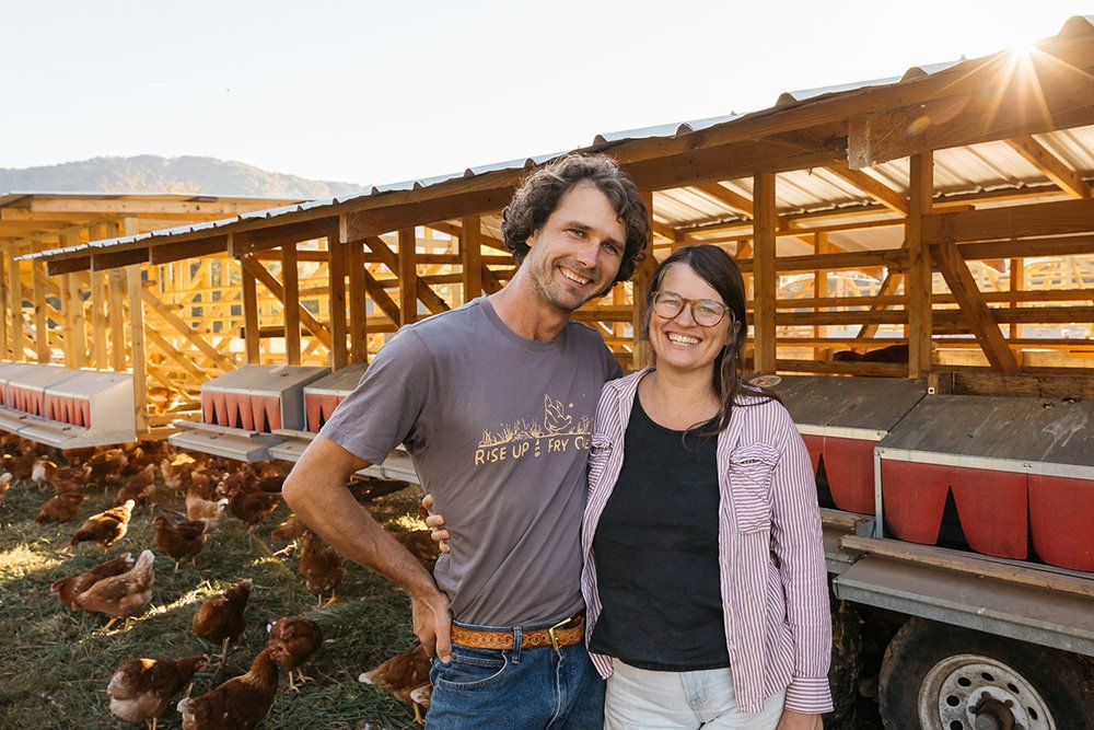 sequatchie.cove.farm.tennessee.pasture.raised.eggs.cover.kelsey.ashley Smiling couple standing together on a poultry farm, surrounded by free-range chickens and wooden chicken coops in the background, with mountains in the distance and sunlight illuminating the scene.